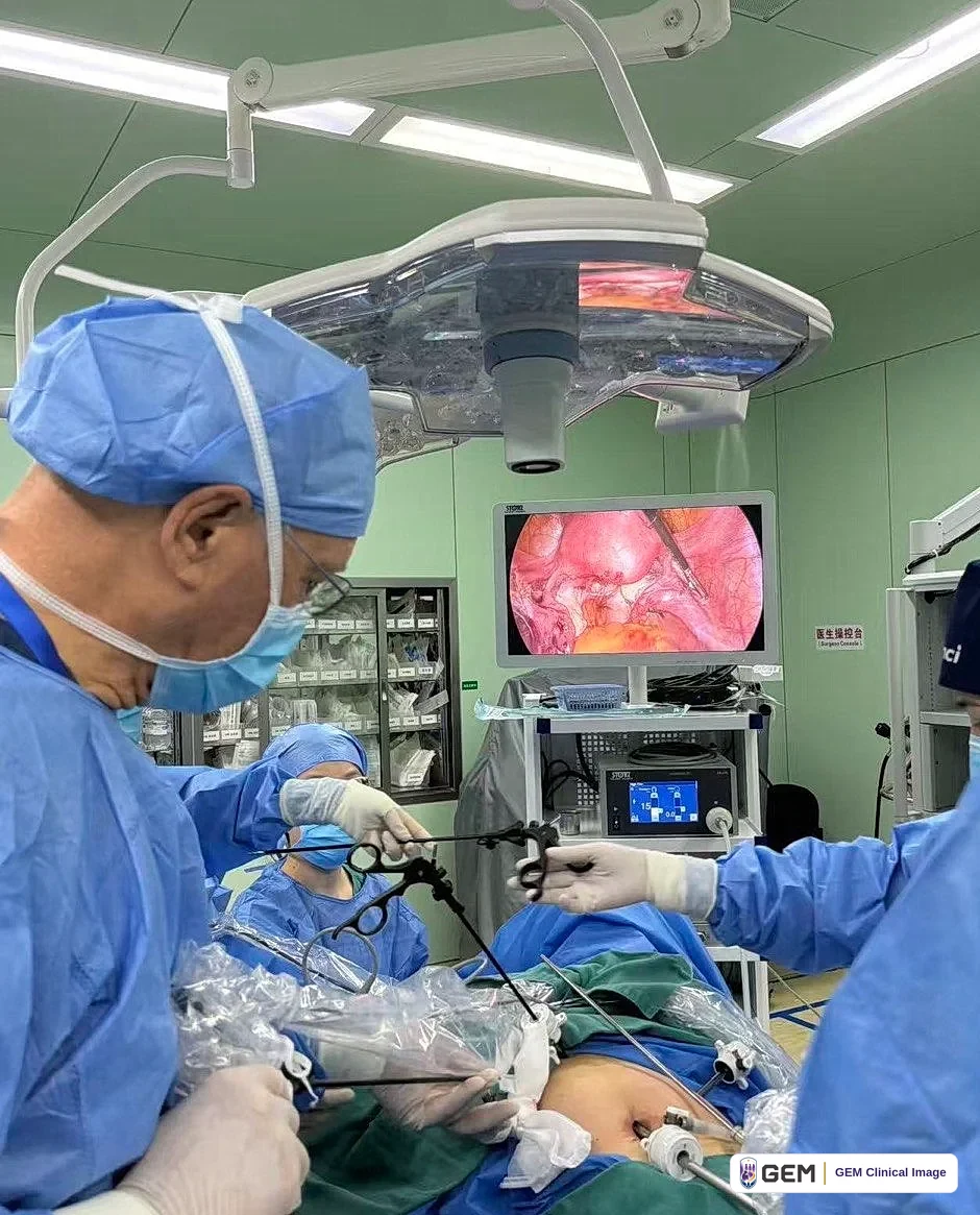 Operating-room photograph: a surgeon in a blue cap, mask and gown holds long laparoscopic instruments over a draped patient, with an overhead surgical light above and a wall monitor showing a laparoscopic view.